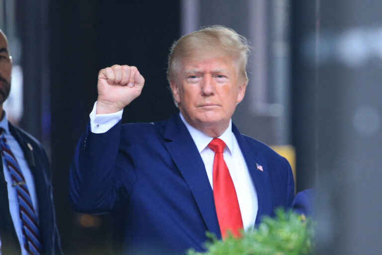 Imagem frontal de Donald Trump, com terno azul e gravata vermelha, com o o braço direito levantado e o punho fechado
