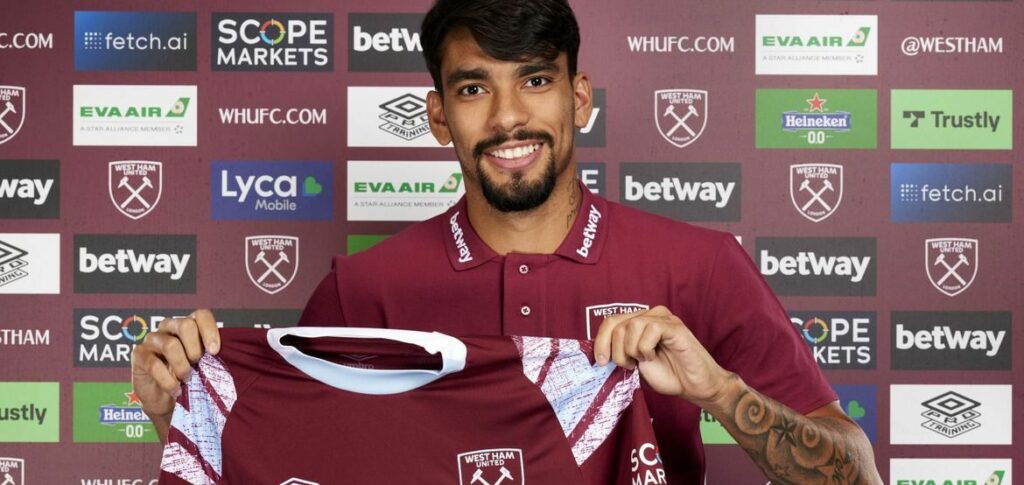 jogador Lucas Paquetá. Homem moreno, com cabelos lisos. Ele está vestindo uma camisa do time, e sorri para foto
